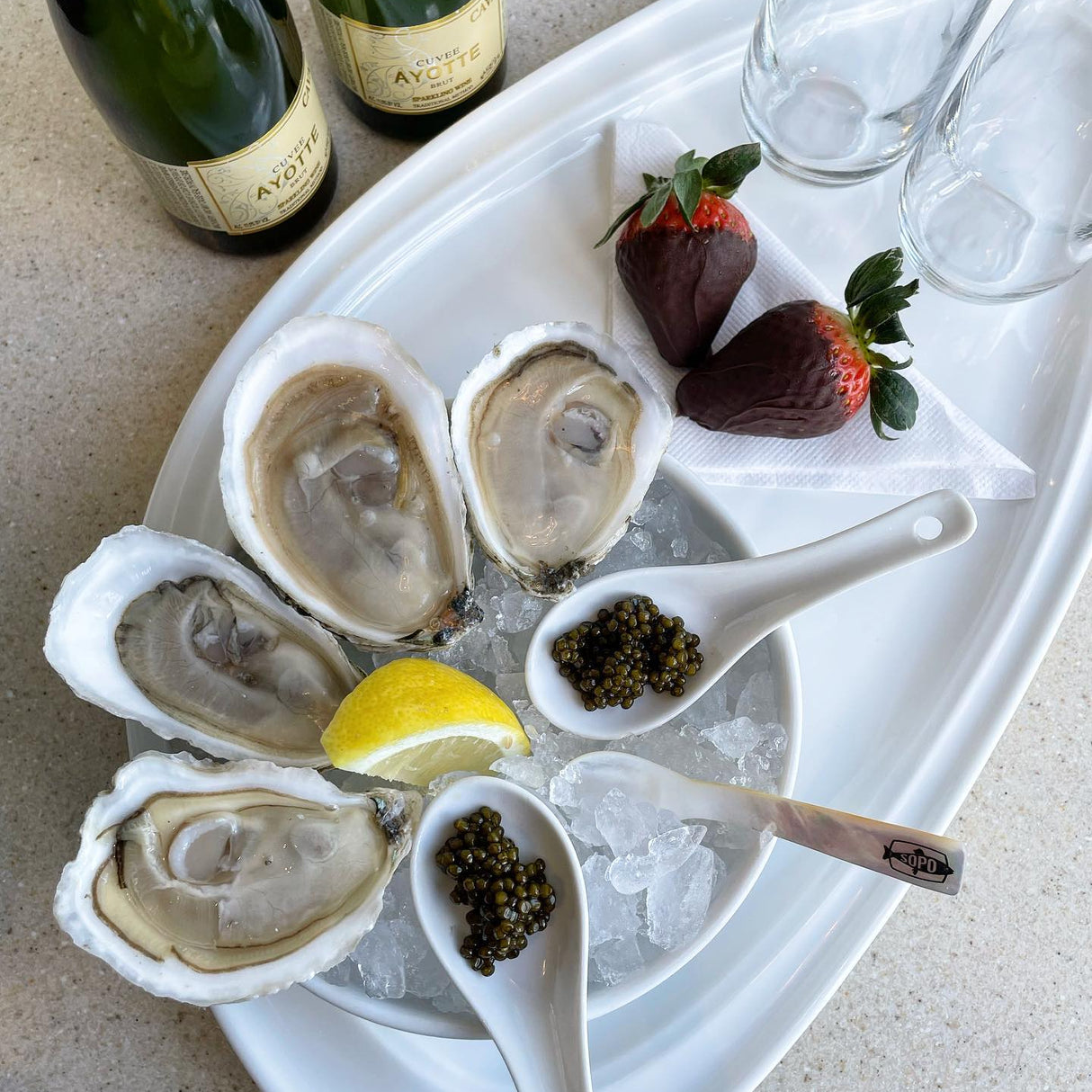 Platter of oysters on ice with caviar, lemon, and chocolate-covered strawberries, accompanied by bottles of champagne.