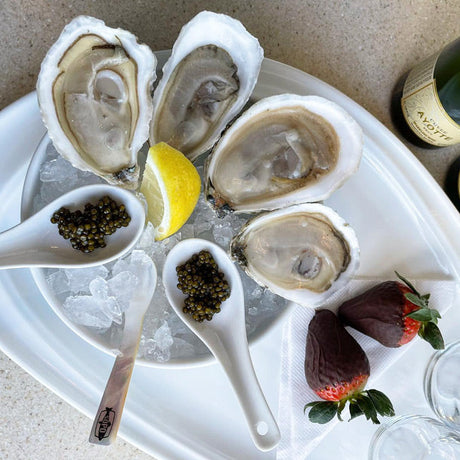 Oysters on a bed of ice with caviar, lemon, and strawberries on a white plate, accompanied by champagne bottles.